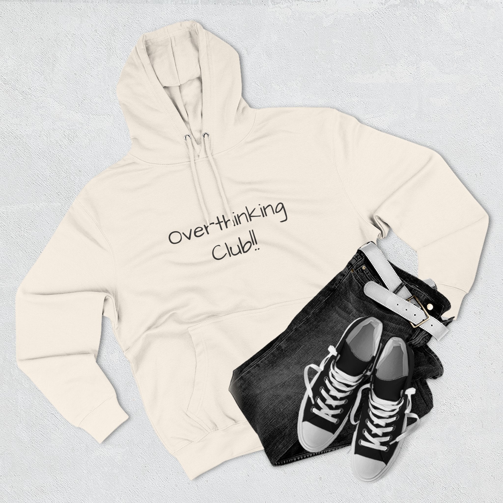 Beige hoodie with 'Overthinking Club!' text, black jeans, and black sneakers on a light gray background